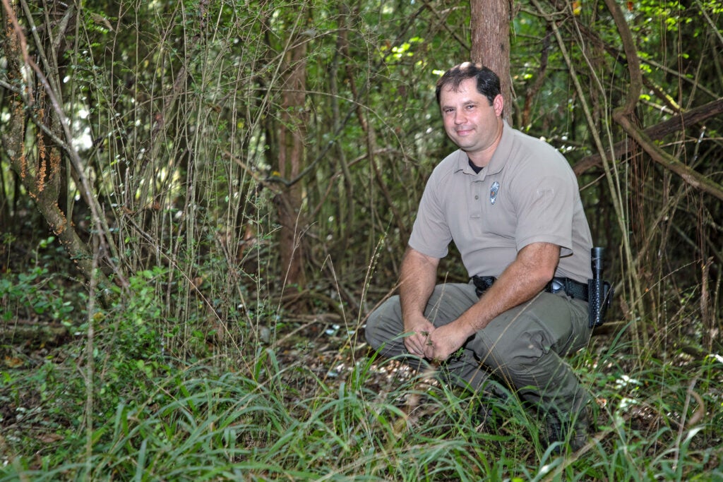 conservation officer crouches in the woods and smiles at camera