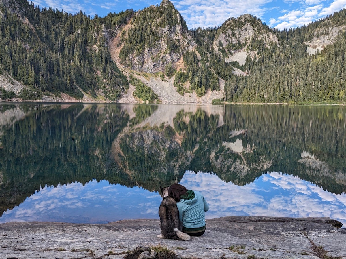 Female hiker wearing Ibex Wool Aire Hoodie sitting on shore of lake with dog