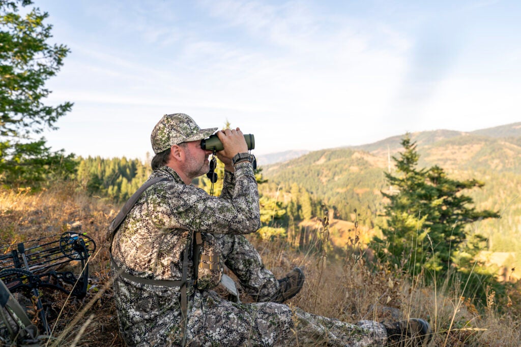 Bowhunter sits on a mountain peak and looks through binoculars while tracking wild game in the forested wilderness of Washington State. A crossbow is lying on the ground behind the man.