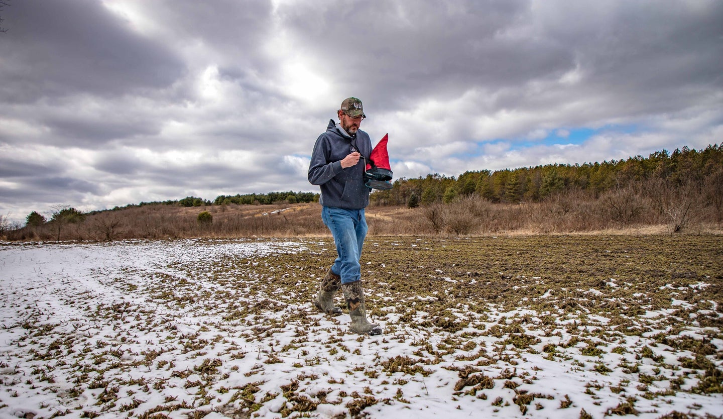 A man walking in a partially snow covered field, spreading clover seed.