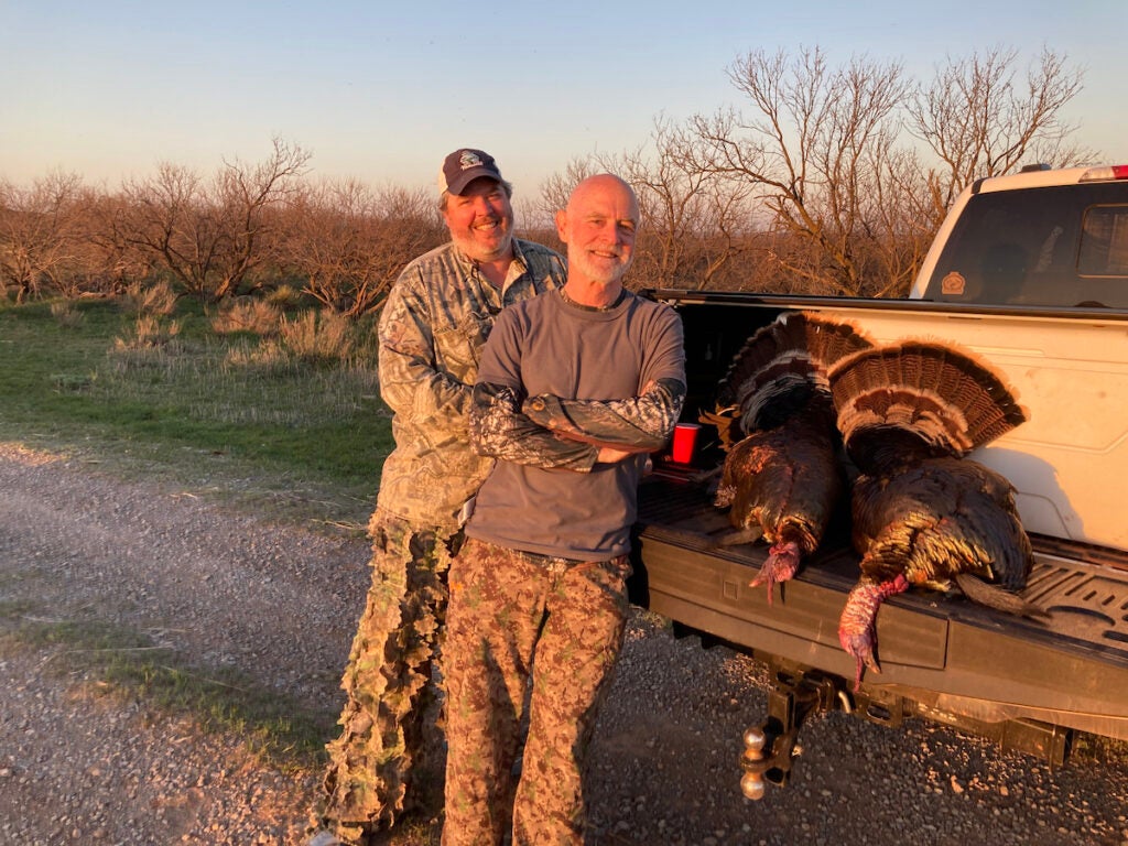 Two men pose next to a pickup truck bed with two dead turkeys.