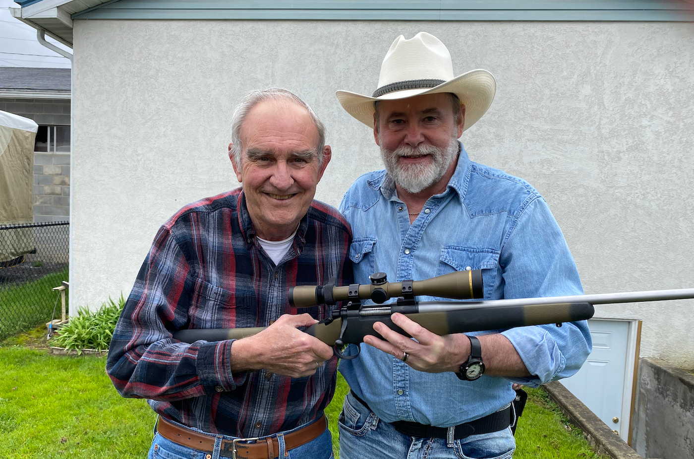 Two men standing in a yard holding a bolt-action rifle.