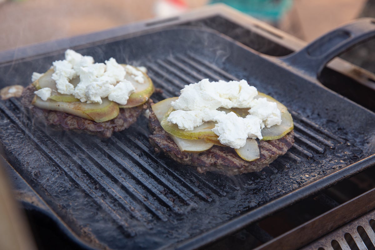 Two burgers on a grill topped with pears and goat cheese.