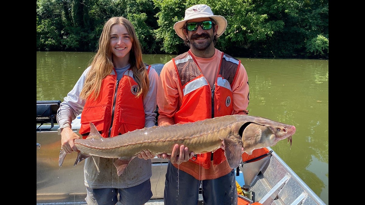 man and woman in lifejackets hold large lake sturgeon