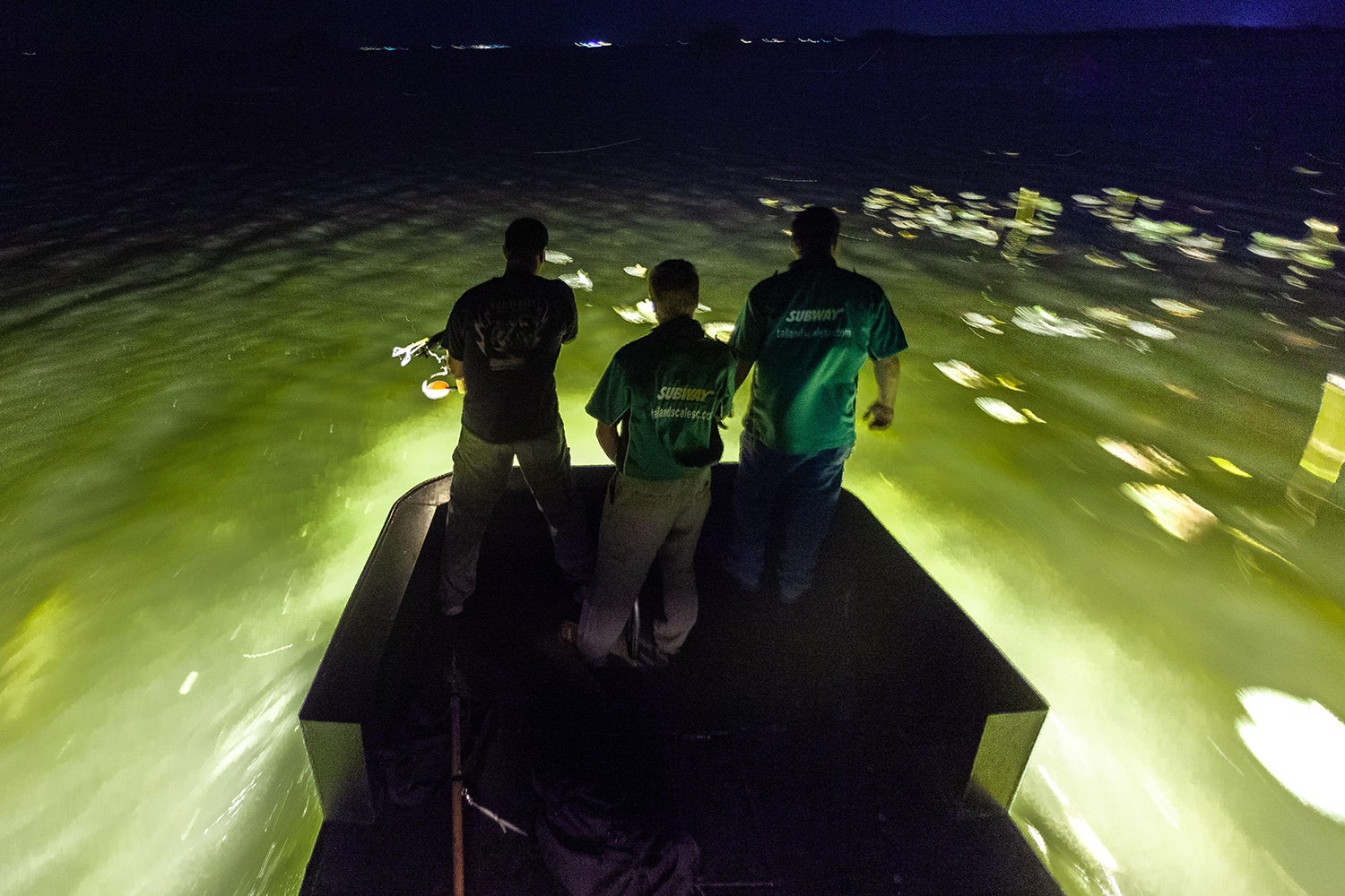 three hunters look out from boat into lit-up water at night