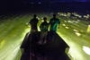 three hunters look out from boat into lit-up water at night