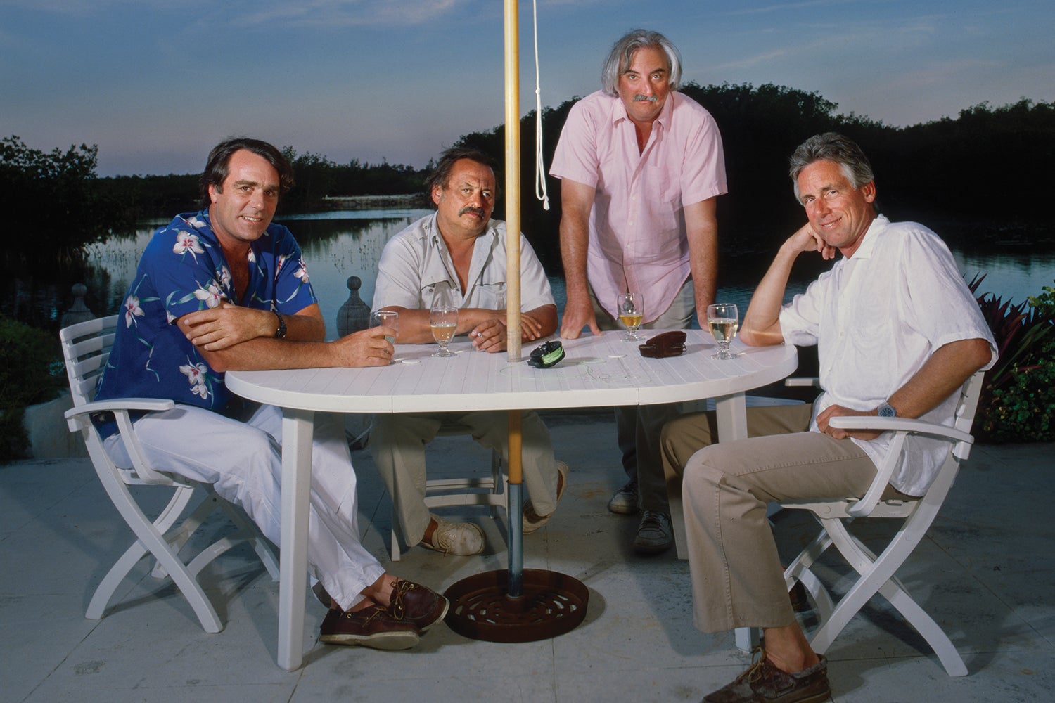 four men with drinks pose around an outdoor table with a creek in the background