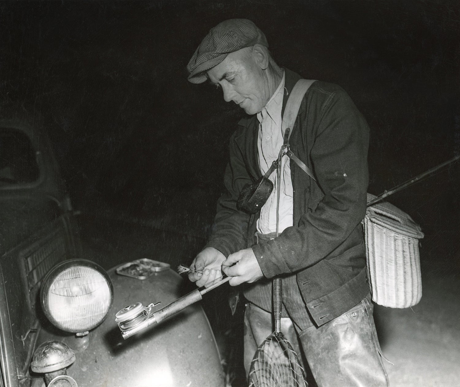 angler wearing wicker creel and holding fly rod ties a fly on his line using his car headlamp for light