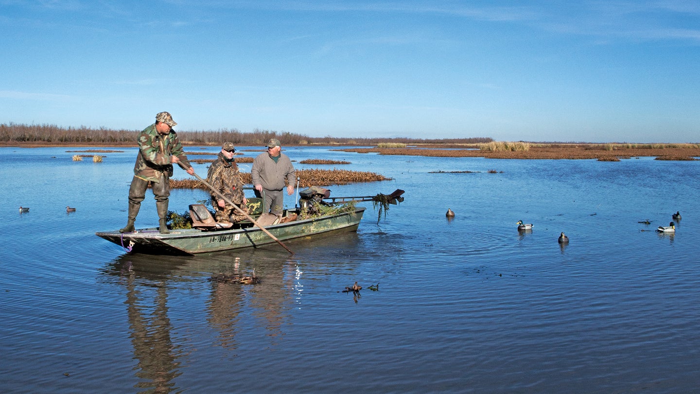 three men wearing camo stand in a small green boat surrounded by a few duck decoys and swampy-looking land in the background