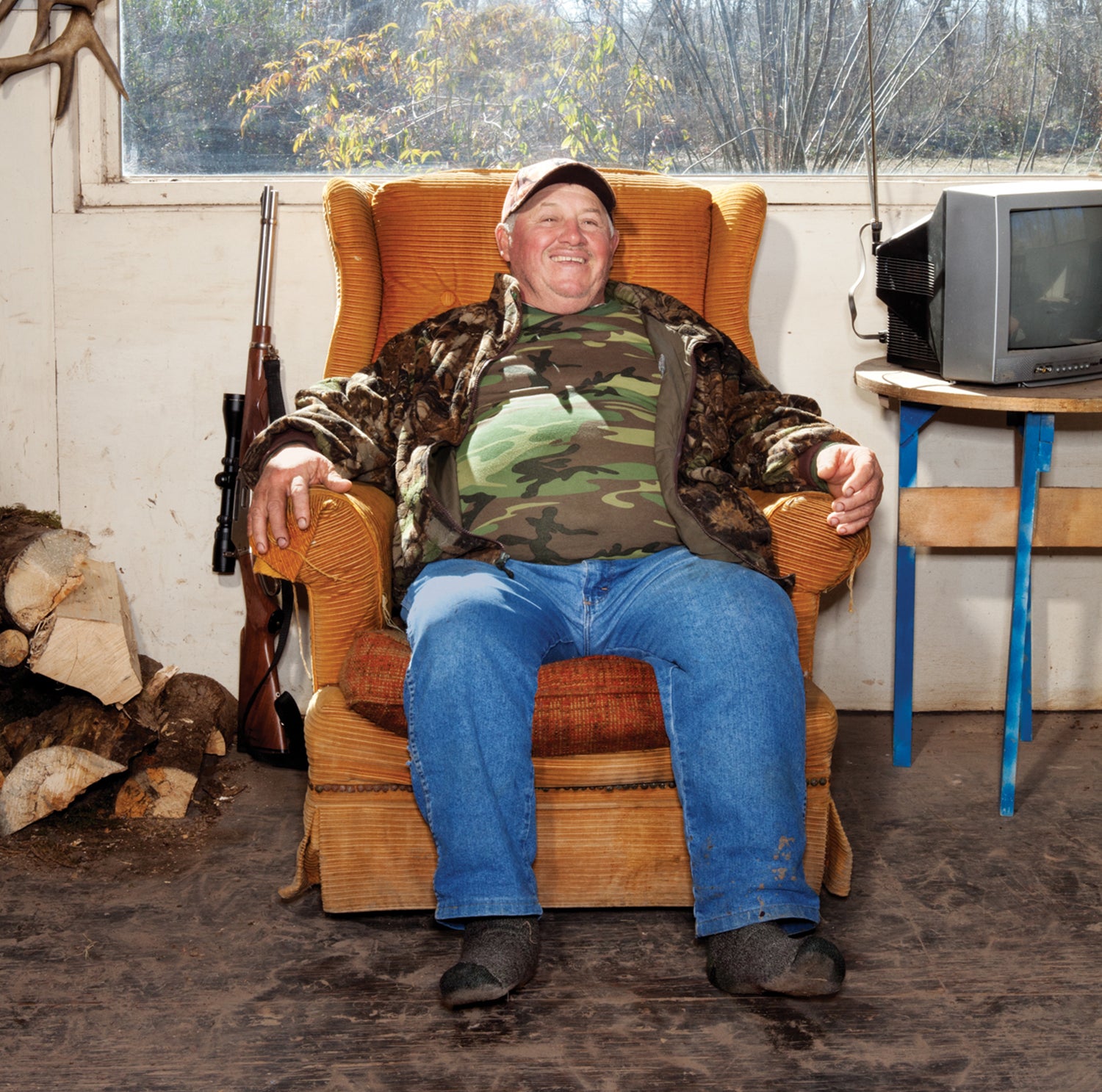 man wearing camo smiles and sits in worn chair under large window