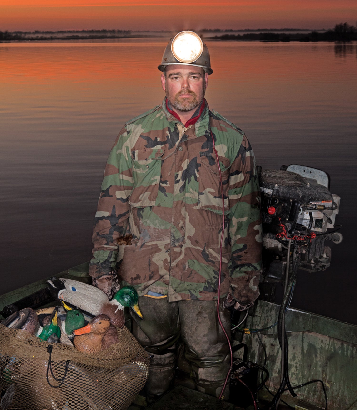 man in camp with large headlamp stands on boat and holds big bag of duck decoys
