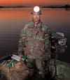 man in camp with large headlamp stands on boat and holds big bag of duck decoys