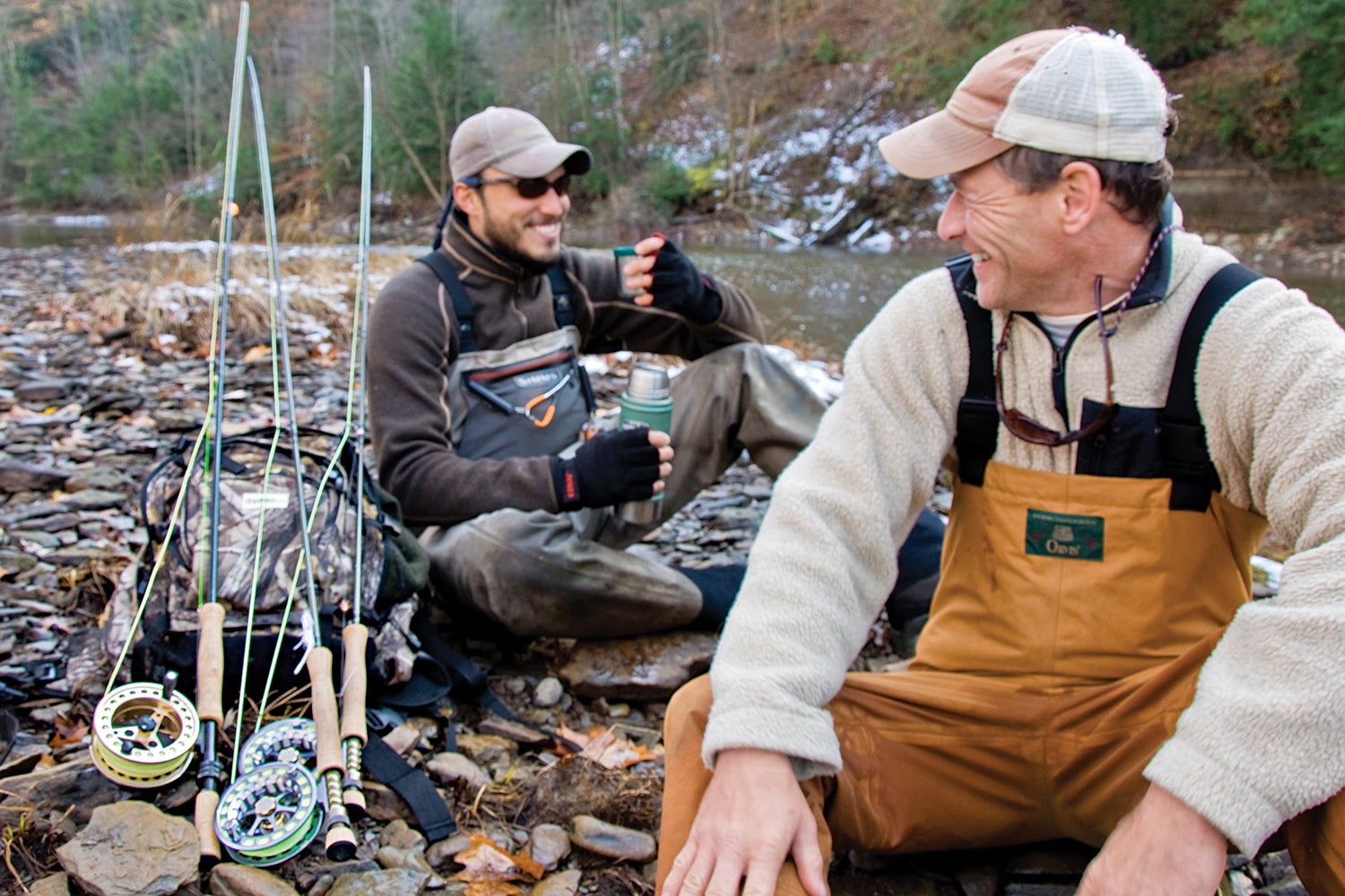 Two anglers rest with a thermost streamside, fly rods beside them.