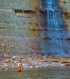 Anglers holding fly rod stands in knee-deep river, waterfall behind him has left bright blue deposits on rock.