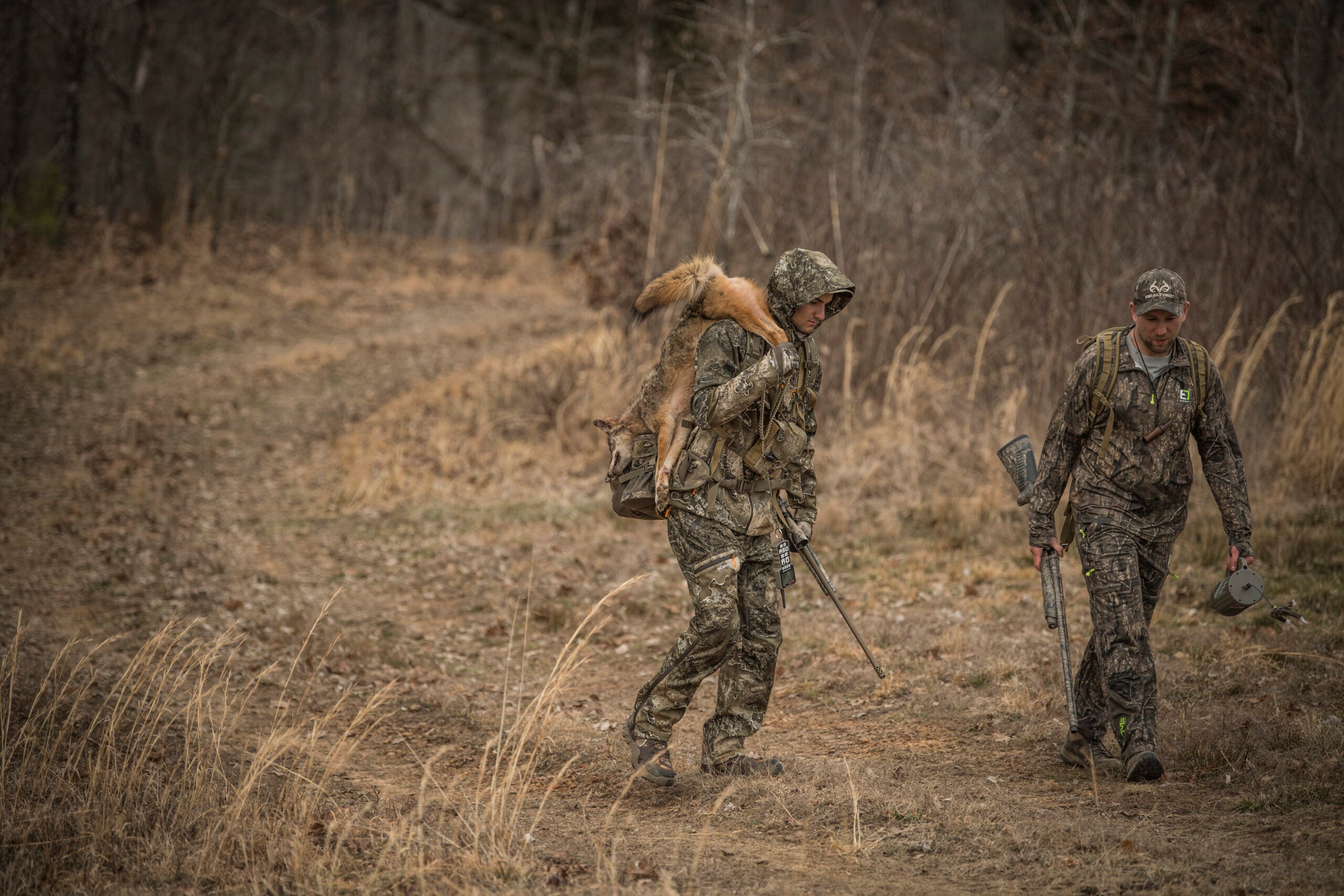 two hunters walk out of the woods, one with a coyote slung over his shoulder
