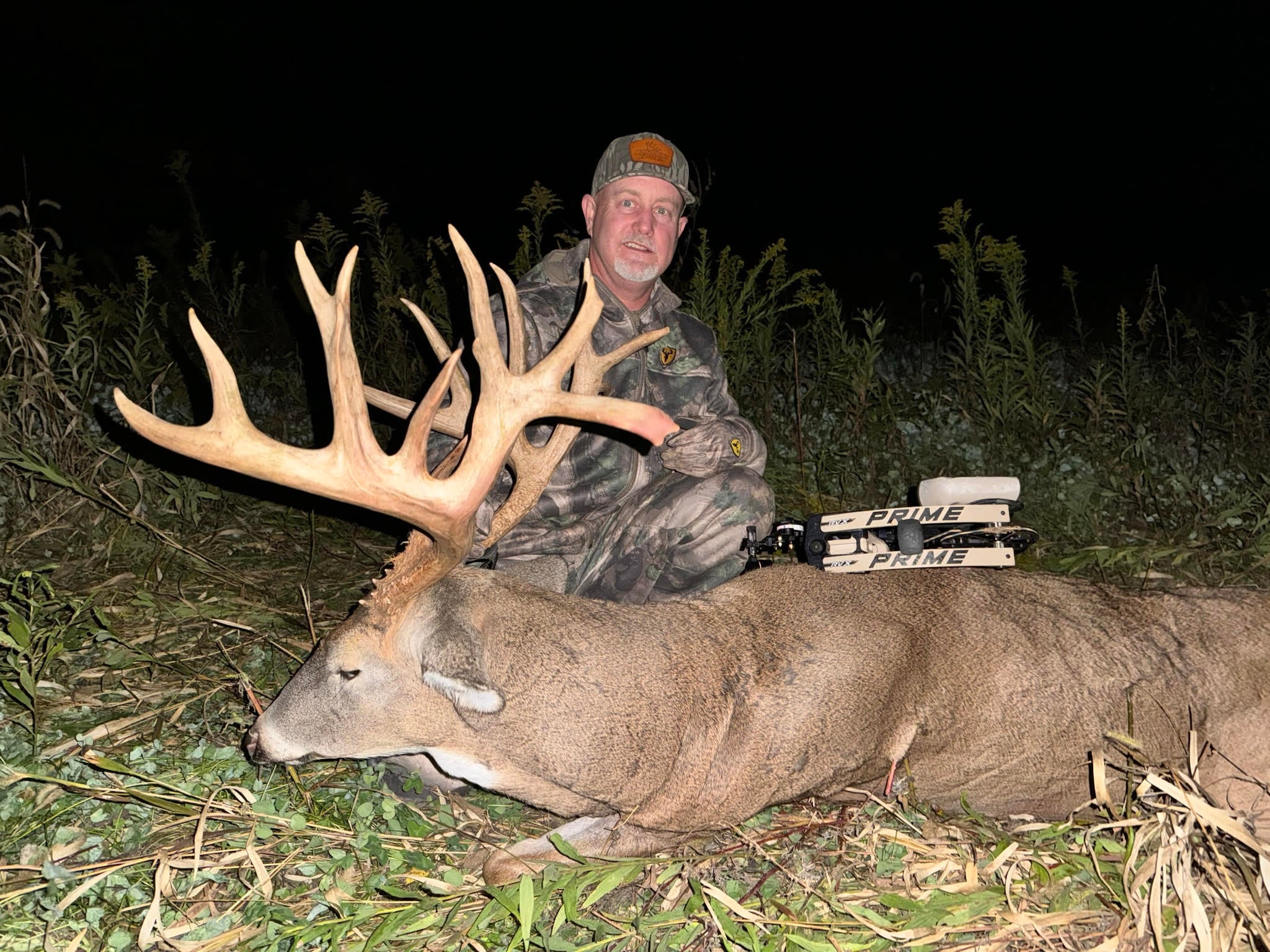 A hunter with a compound bow poses with a huge whitetail buck in a field after dark. 