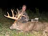 A hunter with a compound bow poses with a huge whitetail buck in a field after dark. 