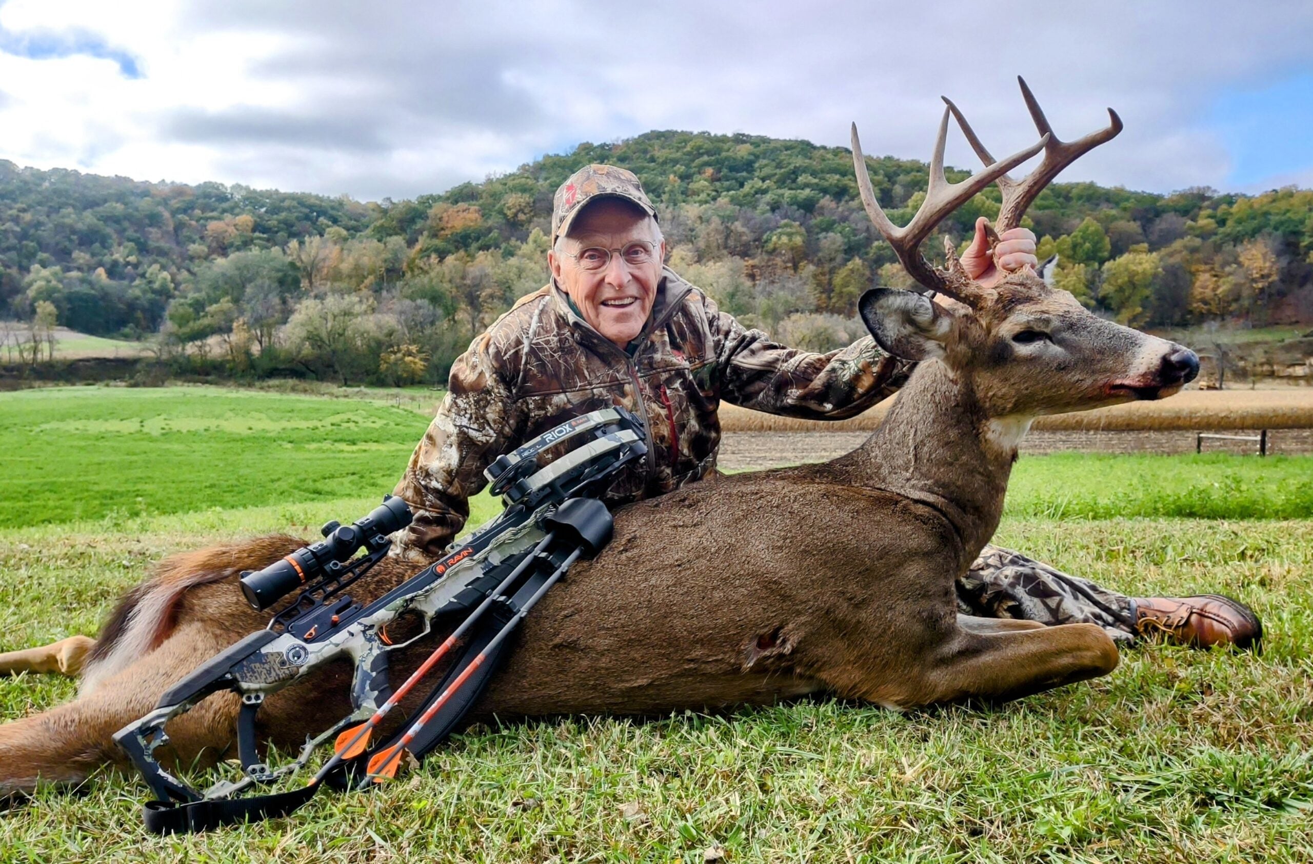 A 93-year-old hunter dressed sits in a green field, posing with a whitetail buck he took with a crossbow.