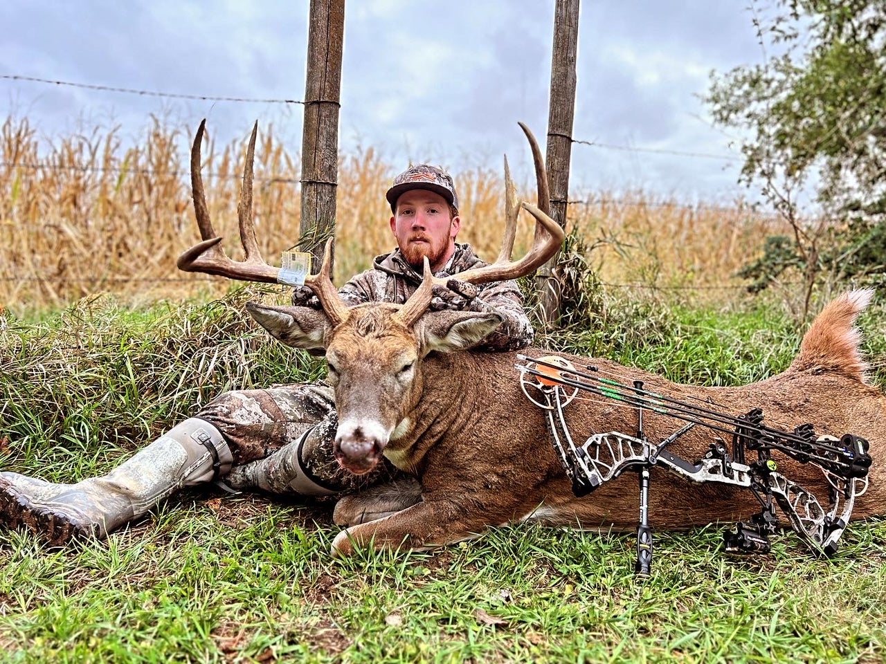 A hunter sits along a wire fence showing off a huge 8-point buck taken with his bow. 