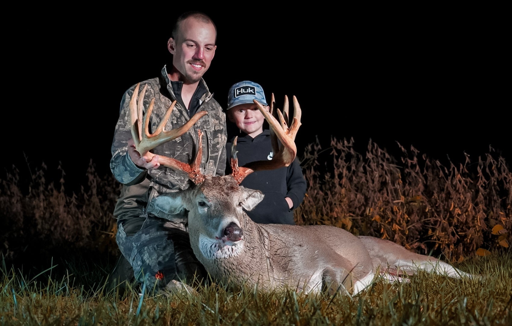 A hunter and his young son sit on the ground, showing off a huge whitetail buck taken with a bow. 