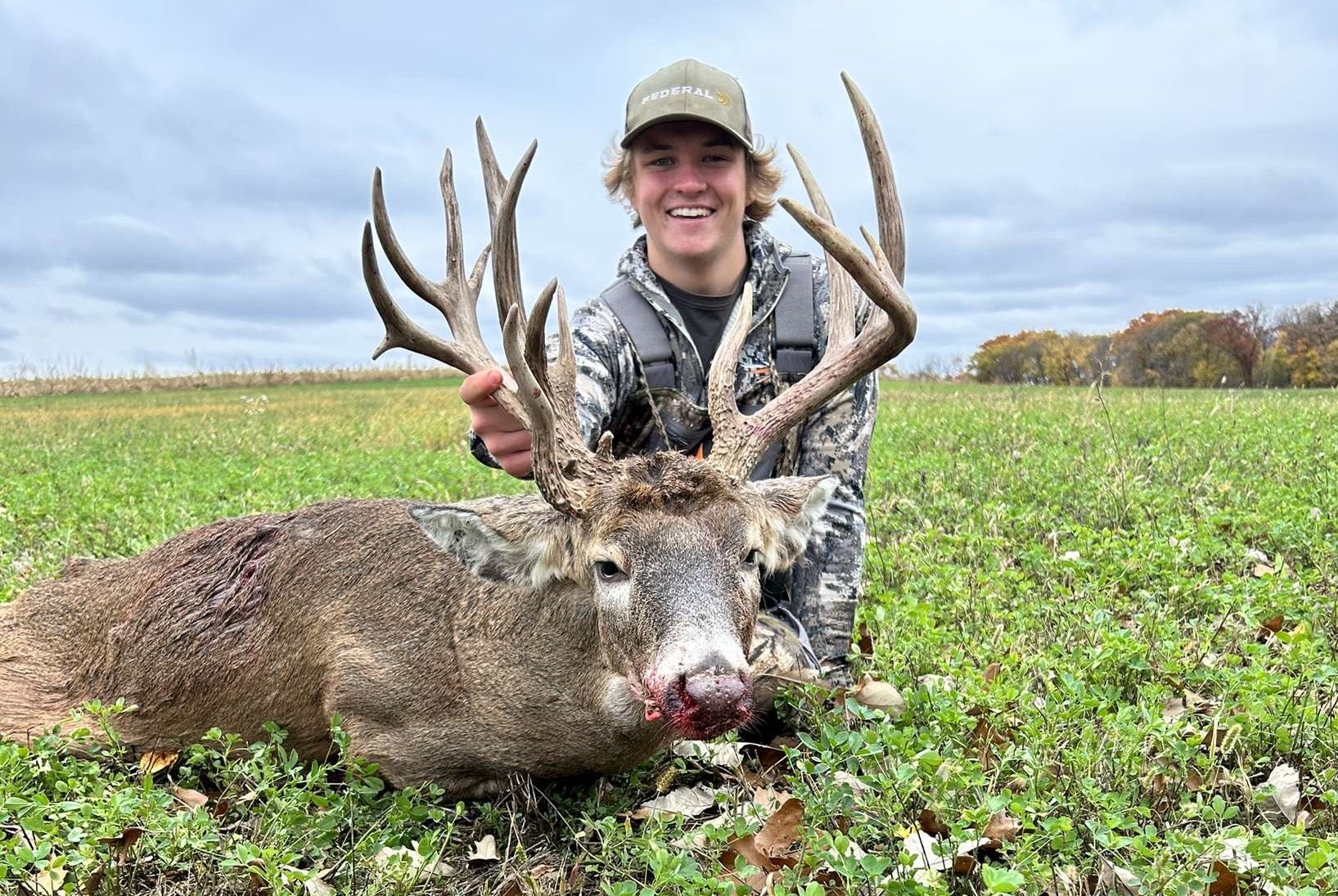 A hunter in a green field sits behind a tall-racked whitetail buck he hunted.