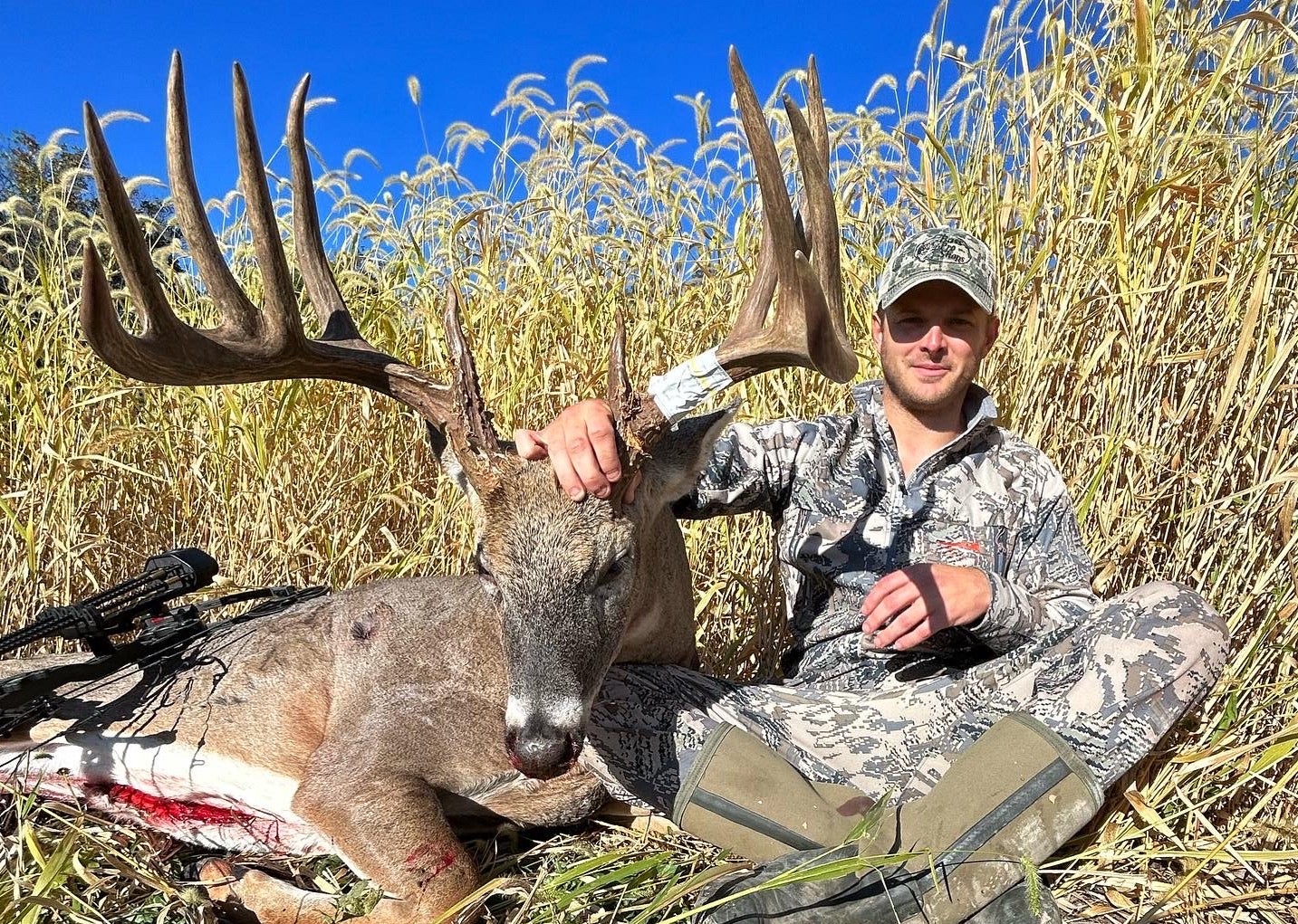 An Iowa hunter sits in field of tall grass, posing with a huge whitetail buck. 