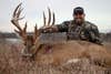 Hunter sits on the ground in a field showing off a huge whitetail buck