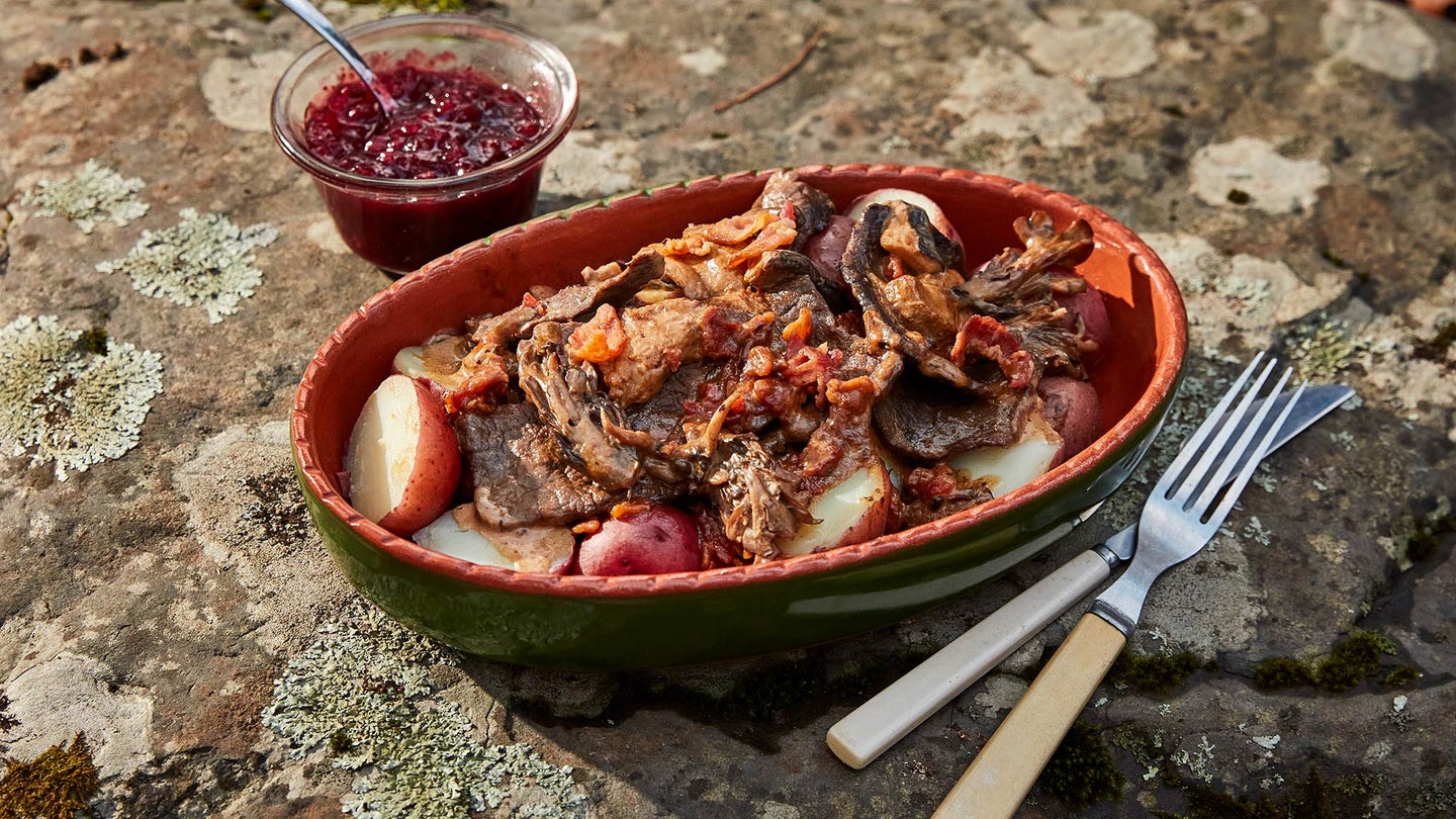 bowl of venison with potatoes and bacon sits on lichen-covered rock along with fork, knife, and small condiment bowl containing red berries