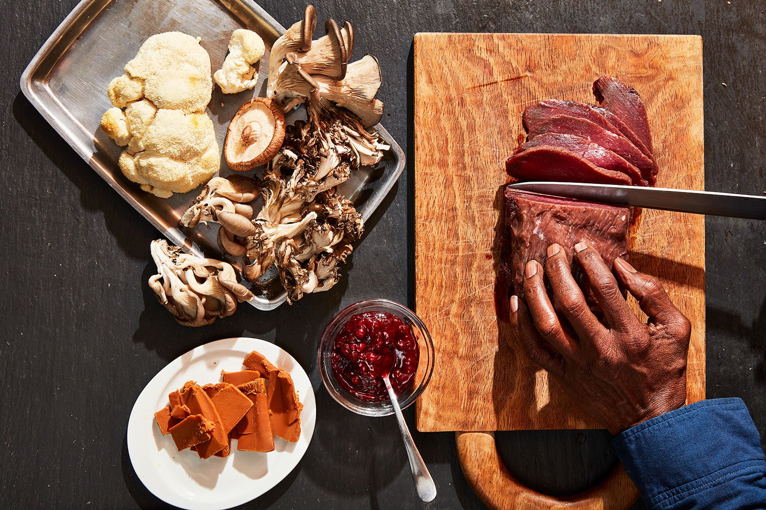 hand holds meat steady as it's being thinly sliced on cutting board sitting next to pay of mushrooms, plate of cheese, and bowl of berries