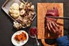 hand holds meat steady as it's being thinly sliced on cutting board sitting next to pay of mushrooms, plate of cheese, and bowl of berries