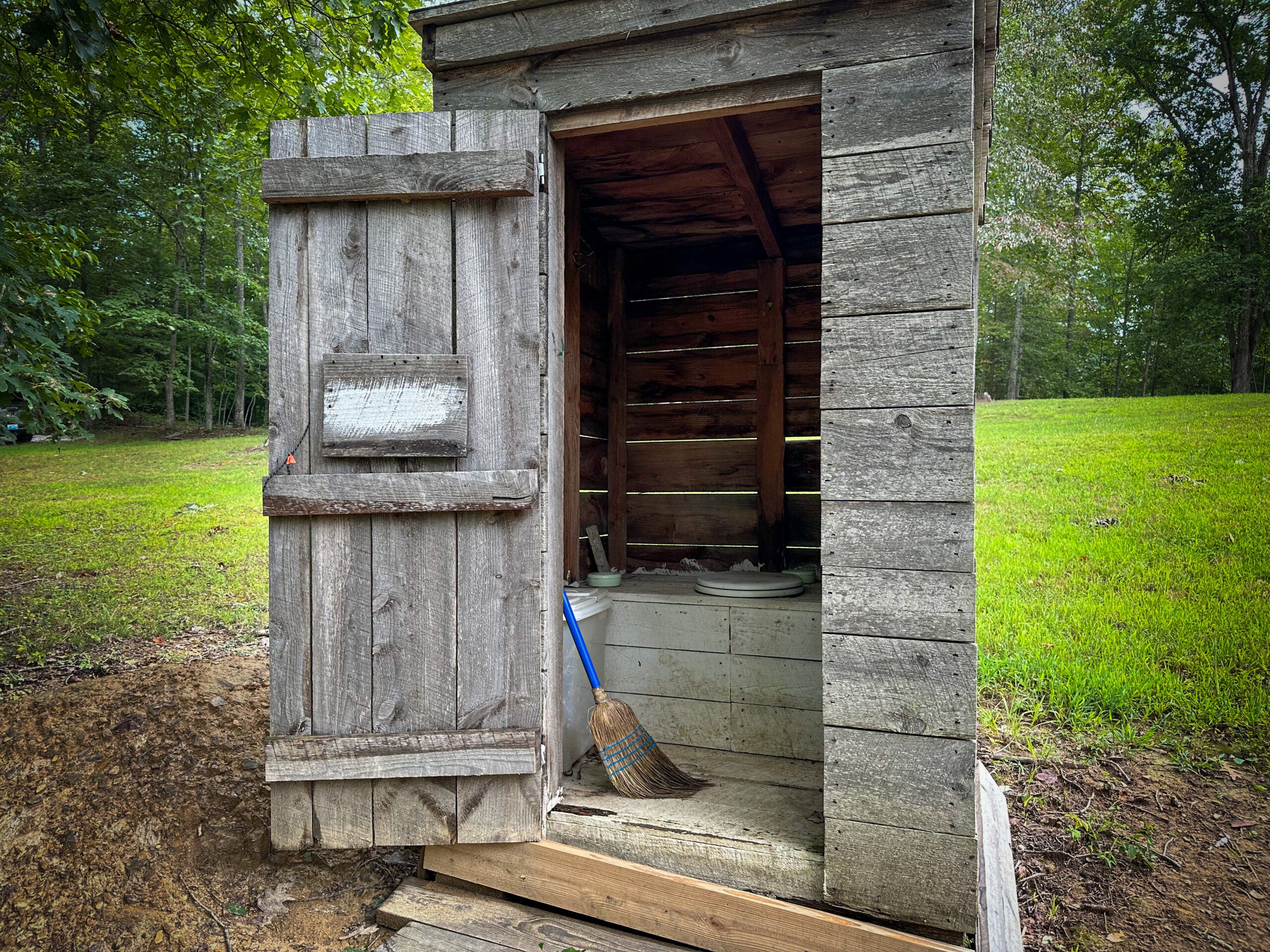 A small wooden outhouse with the door open, in front of a patch of green grass.