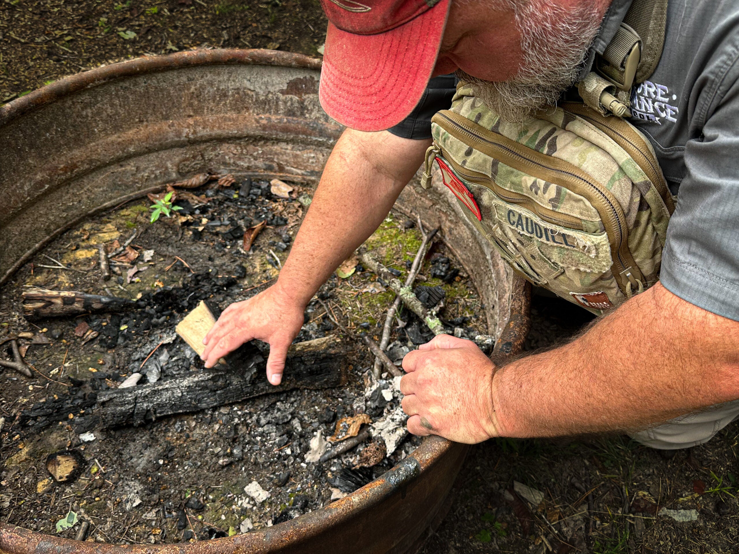 A camper wearing a red hat and a camo chest pack leans over a circular fire pit in the woods to make sure the coals are not smoldering.