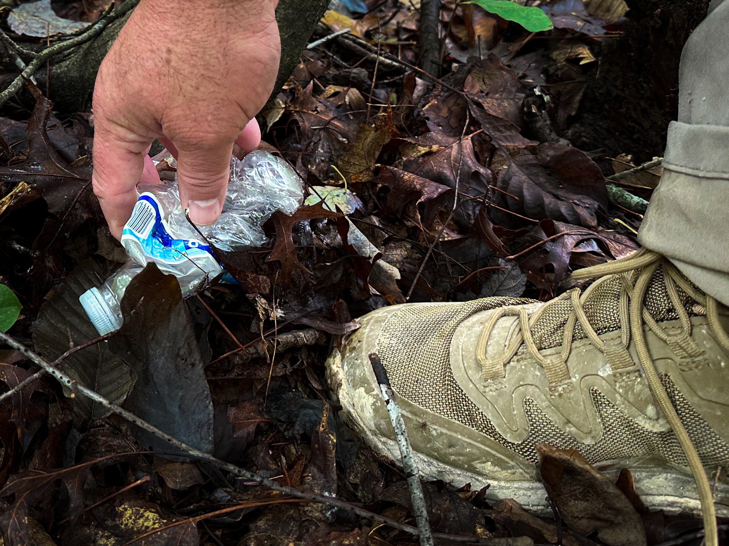 A camper picks up a piece of trash on the ground in the woods that is next to his hiking boot.