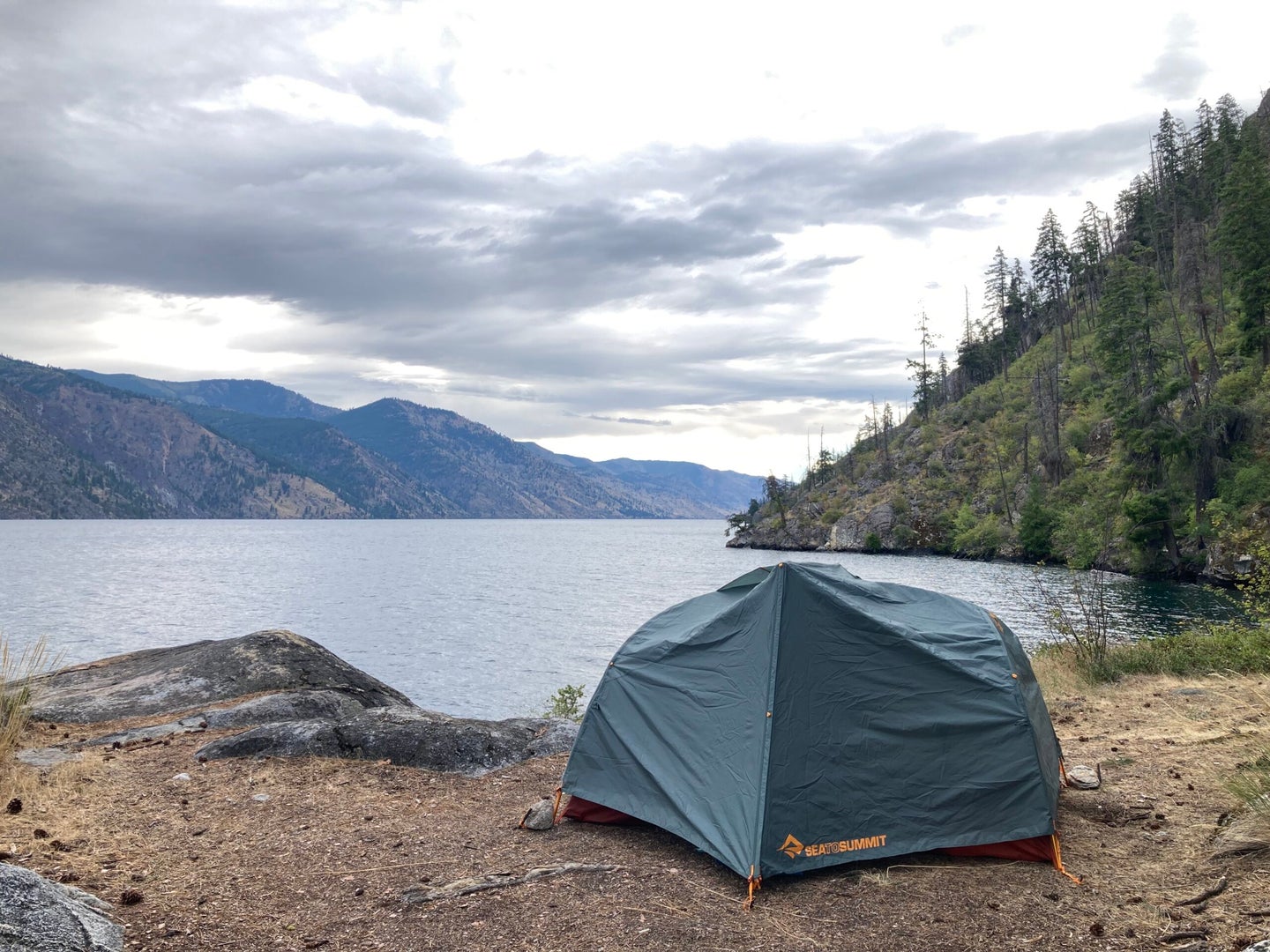 A green camping tent pitched on the ground in front of a lake and mountains.