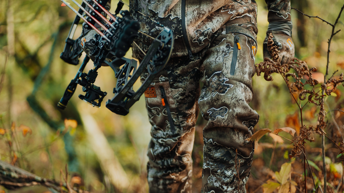 Hunter walking through woods in Scentlok pants holding compound bow