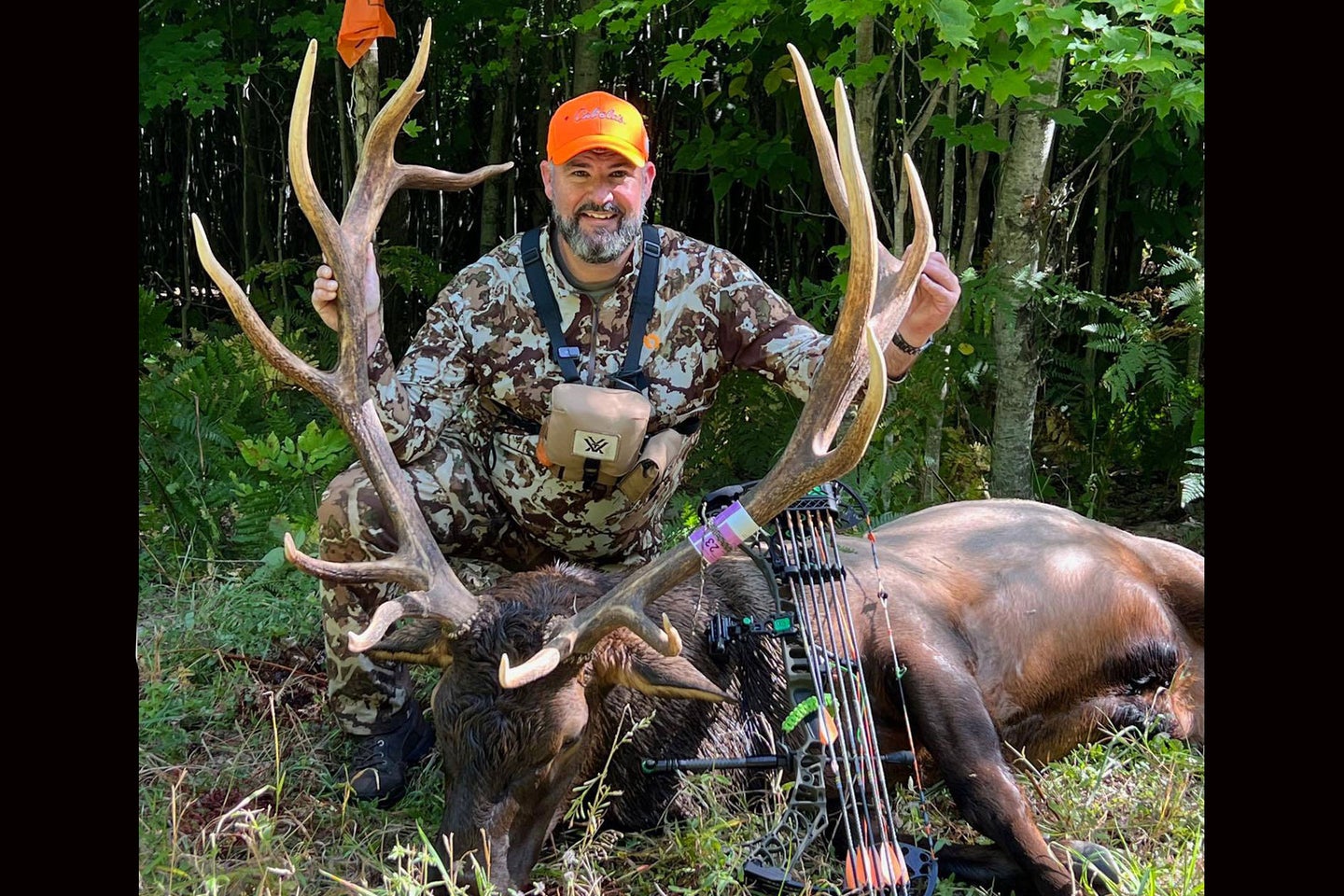 A hunter poses with a new state record bull elk against a forested backdrop.