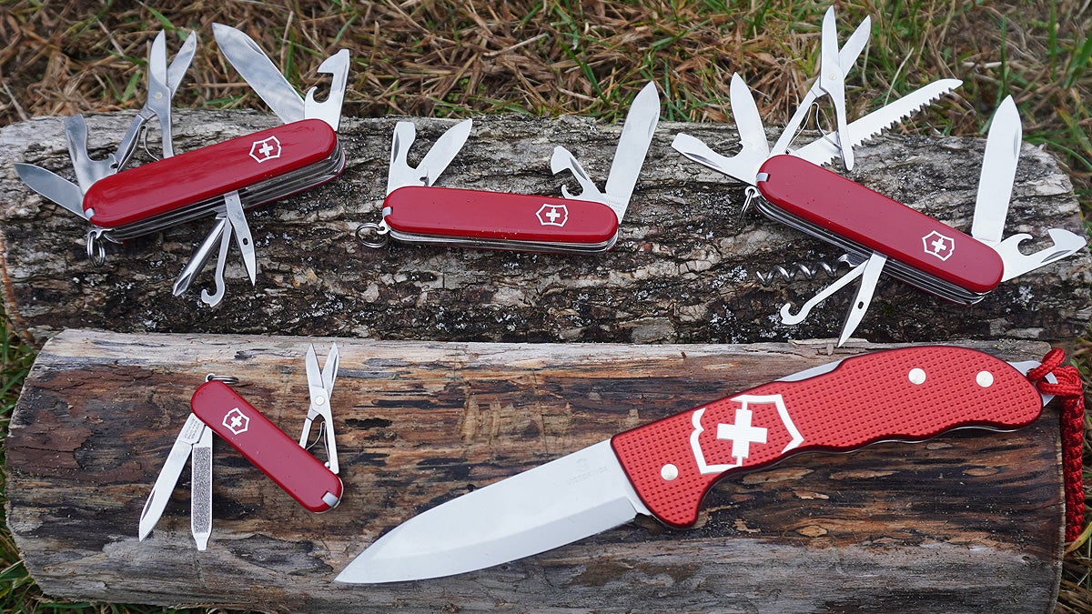 Five red and silver Swiss Army branded knives with various tools unfolded sitting on two wet logs with and without bark.