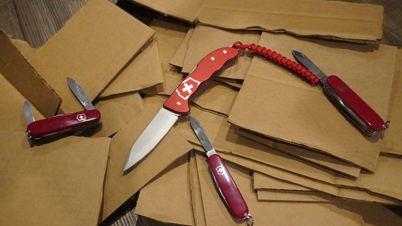 Four red and silver Swiss Army knives with blades extended sitting on a pile of sliced up brown cardboard. 