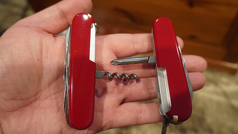 Two red and white Swiss Army knives with the corkscrew and screwdriver tools extended on the palm of a hand. 