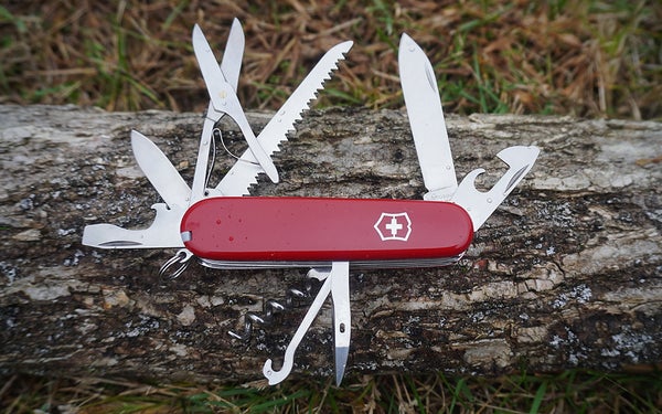 A red and white Victorinox Swiss Army Huntsman knife with tools unfolded on a grey and white log on grass.