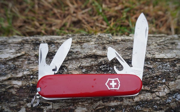 A Victorinox Swiss Army Recruit knife with features unfolded on a grey log sitting on the grass.