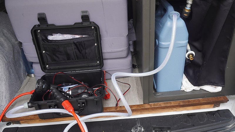 The black Klymit WaterPod shower system case sitting in the back of a tiny van build with a hose running into a blue water tank. 