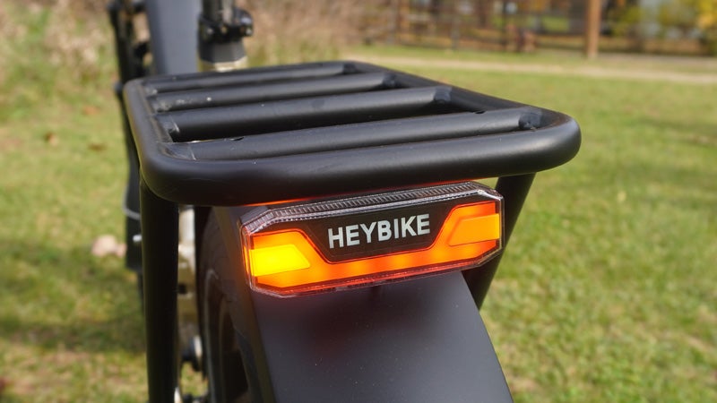 The orange taillight and turn signal of a Heybike electric bicycle below the black luggage rack on a green lawn. 