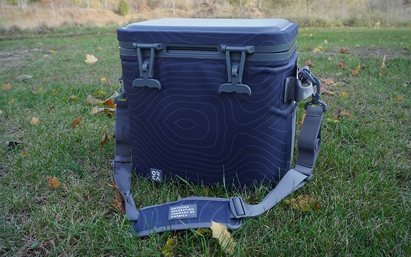 The blue and gray Orca Wanderer 24 cooler with shoulder strap sitting on green grass.