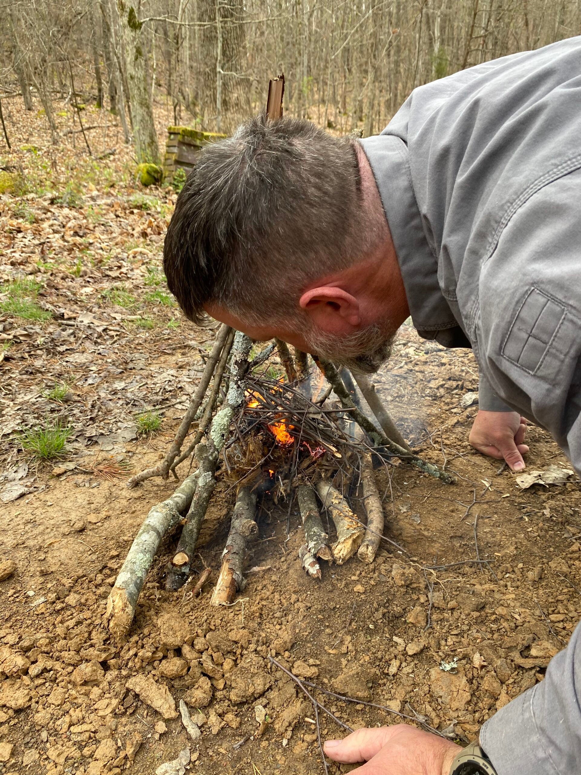 A man leans down to the ground to blow into a campfire to help the flames grow.