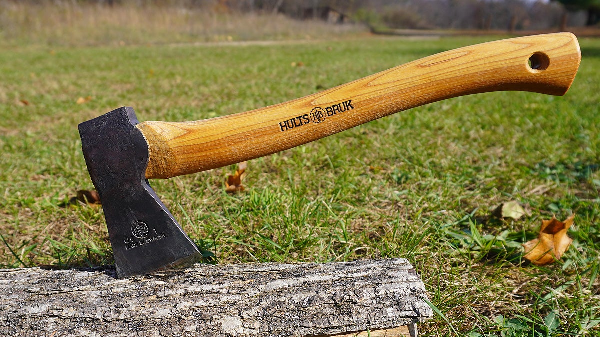 A small Hults Bruk axe with a wooden handle and black axe head in a log on a grassy lawn.
