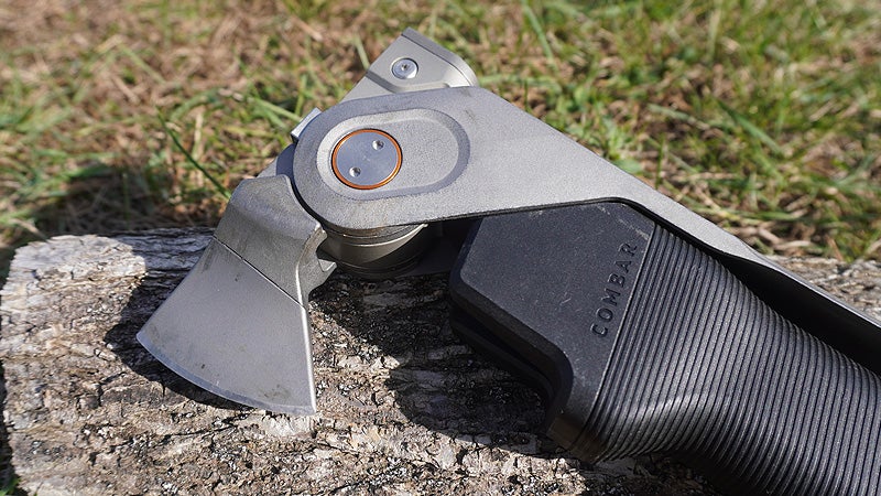 The silver head and black handle of a Combar multi-tool on a log in a grassy lawn. 