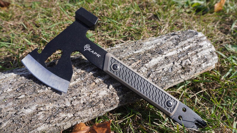 The black and tan Reapr Versa Camp axe leaning against a log on a grassy lawn. 