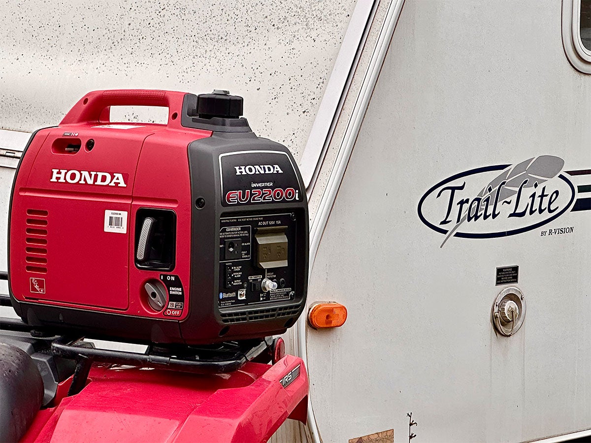 A Honda generator sitting on the rack of a Honda ATV next to a white camper trailer.