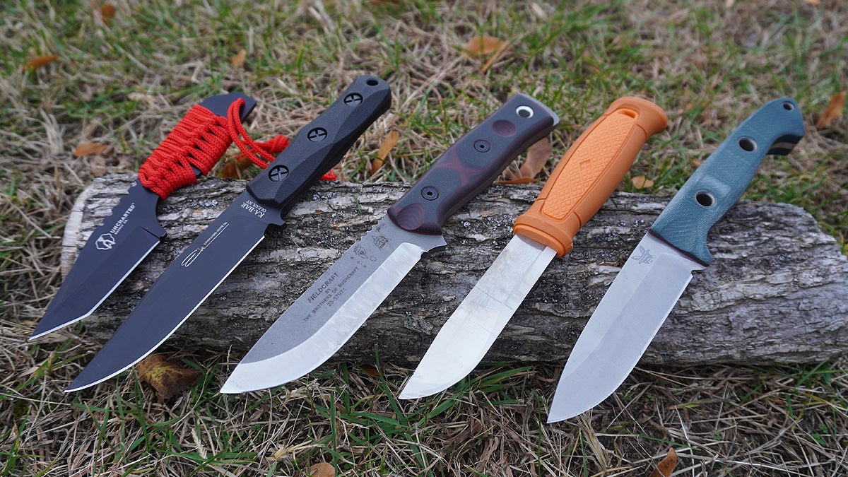Five fixed blade bushcraft ready knives sitting on a log on a grassy lawn.